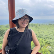 A potrait of Melissa, a non-binary person of Chinese descent with tan skin wearing a bucket hat and black shirt in front of a scenic landscape with greenery.